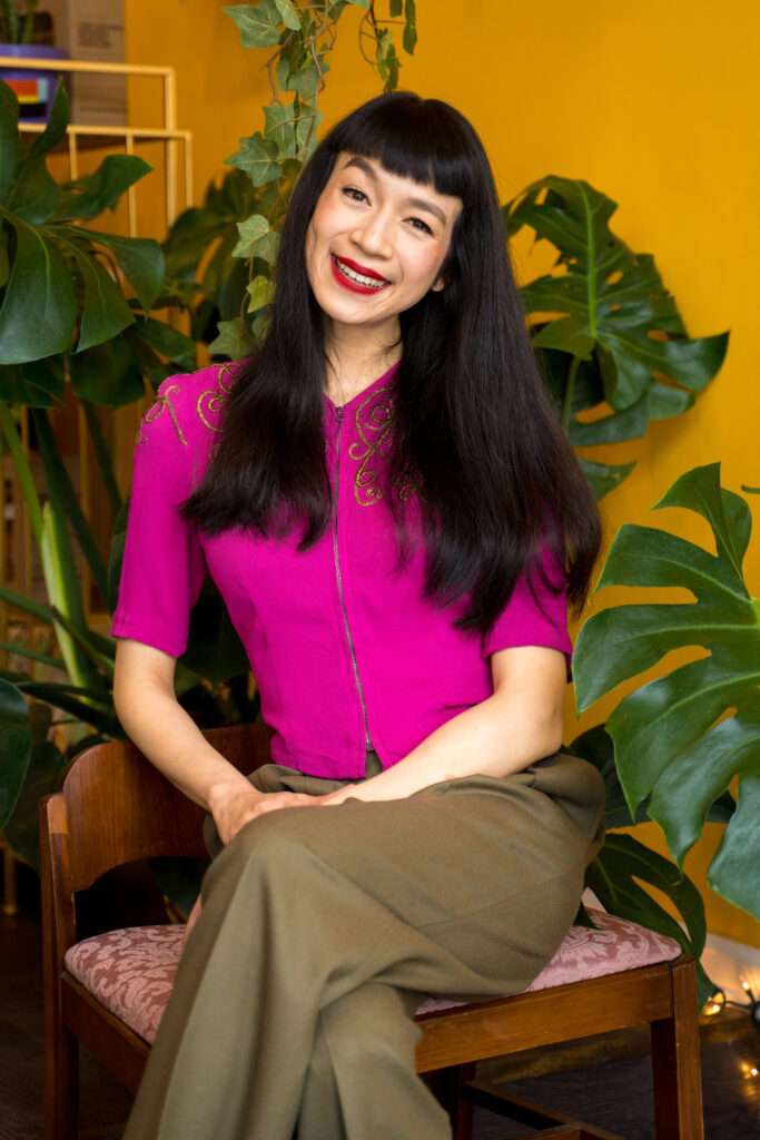 nat vikitsreth of come back to care, chinese-thai woman with long black hair, red lipstick, smiling with head tilted and hands gently folded in lap